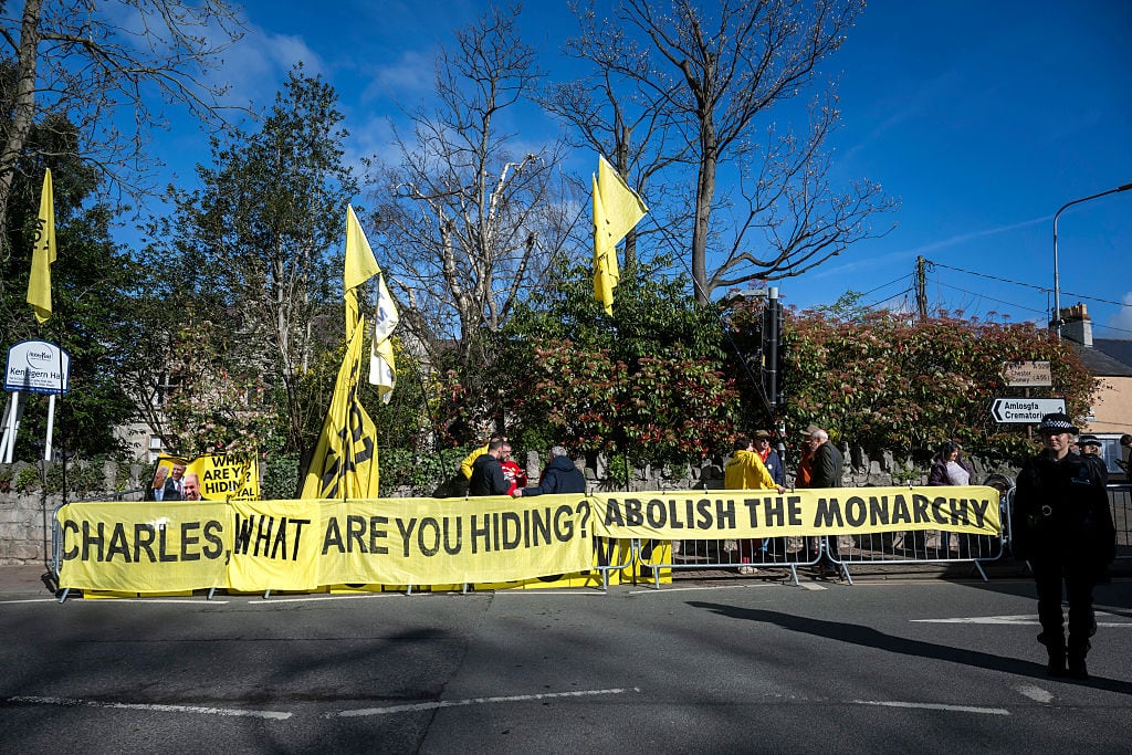 Banners reading "Charles, what are you hiding?" and "Abolish the monarchy" hang from barricades along a street with people standing behind them.