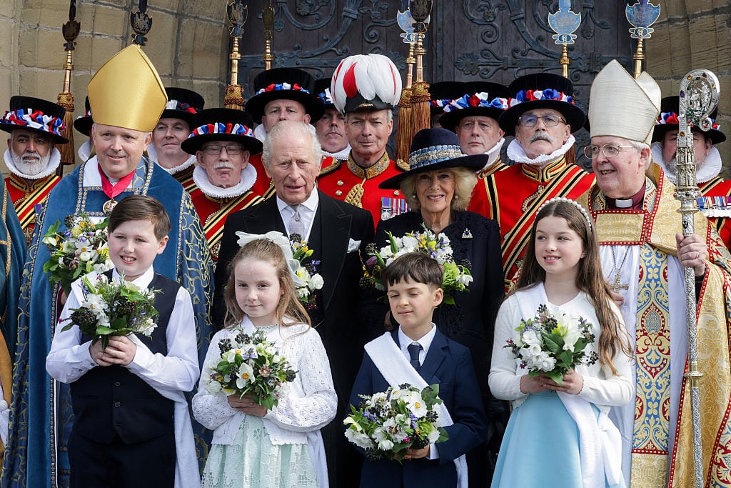 Children and adults holding flowers, along with people in religious vestments and formal military attire stand outside a church.