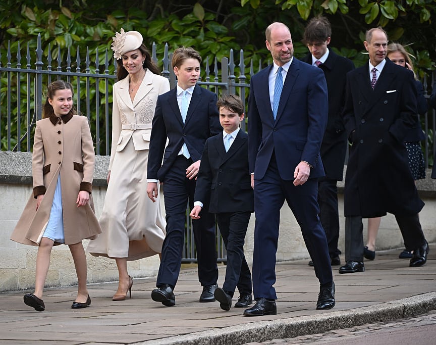 Several children and adults walk along a stone sidewalk beside a high iron fence with trees behind it.