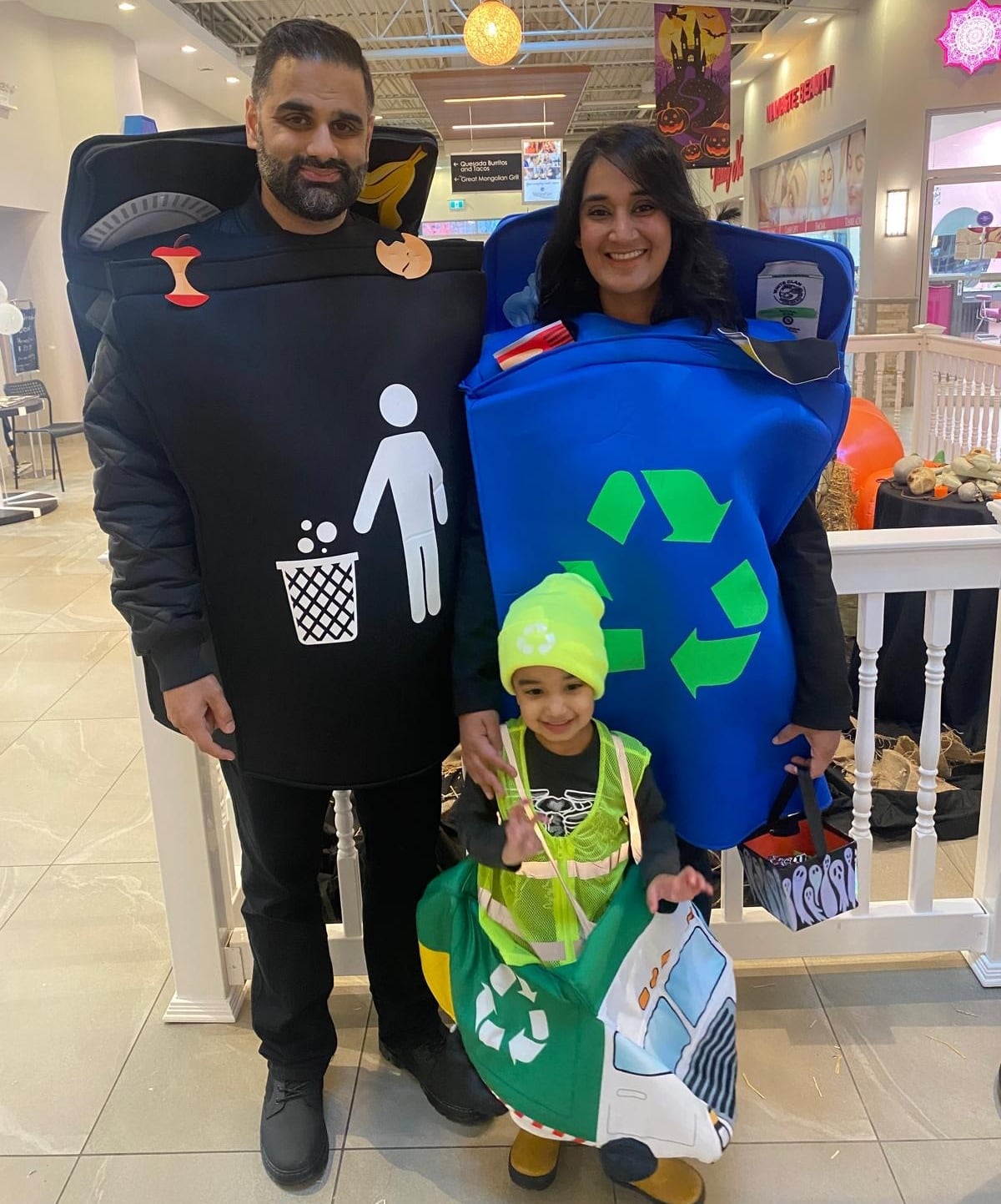 A man, woman and child are seen with recycling-themed costumes. The man is wearing a black shirt with the logo of a man throwing refuse, the woman with a large recycling logo, and the child with a high-vis vest.