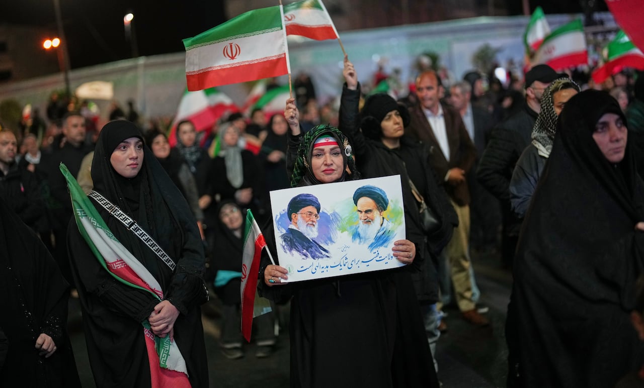 A woman wearing a black chador holds a poster depicting Iran's Ayatollah Ali Khamenei and Ayatollah Ruhollah Khomeini, at night surrounded by a crowd.