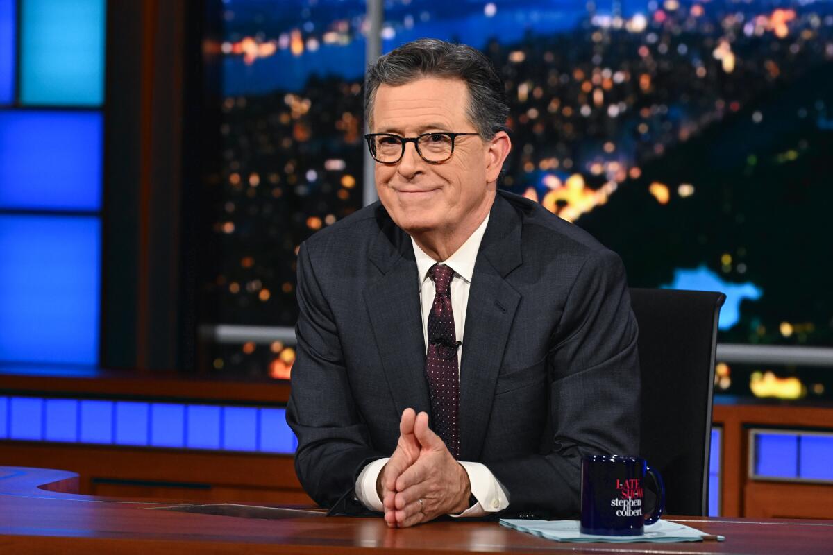 A man with dark hair and glasses, in dark suit and suit, smiles while seated at a desk, his palms together, near a mug