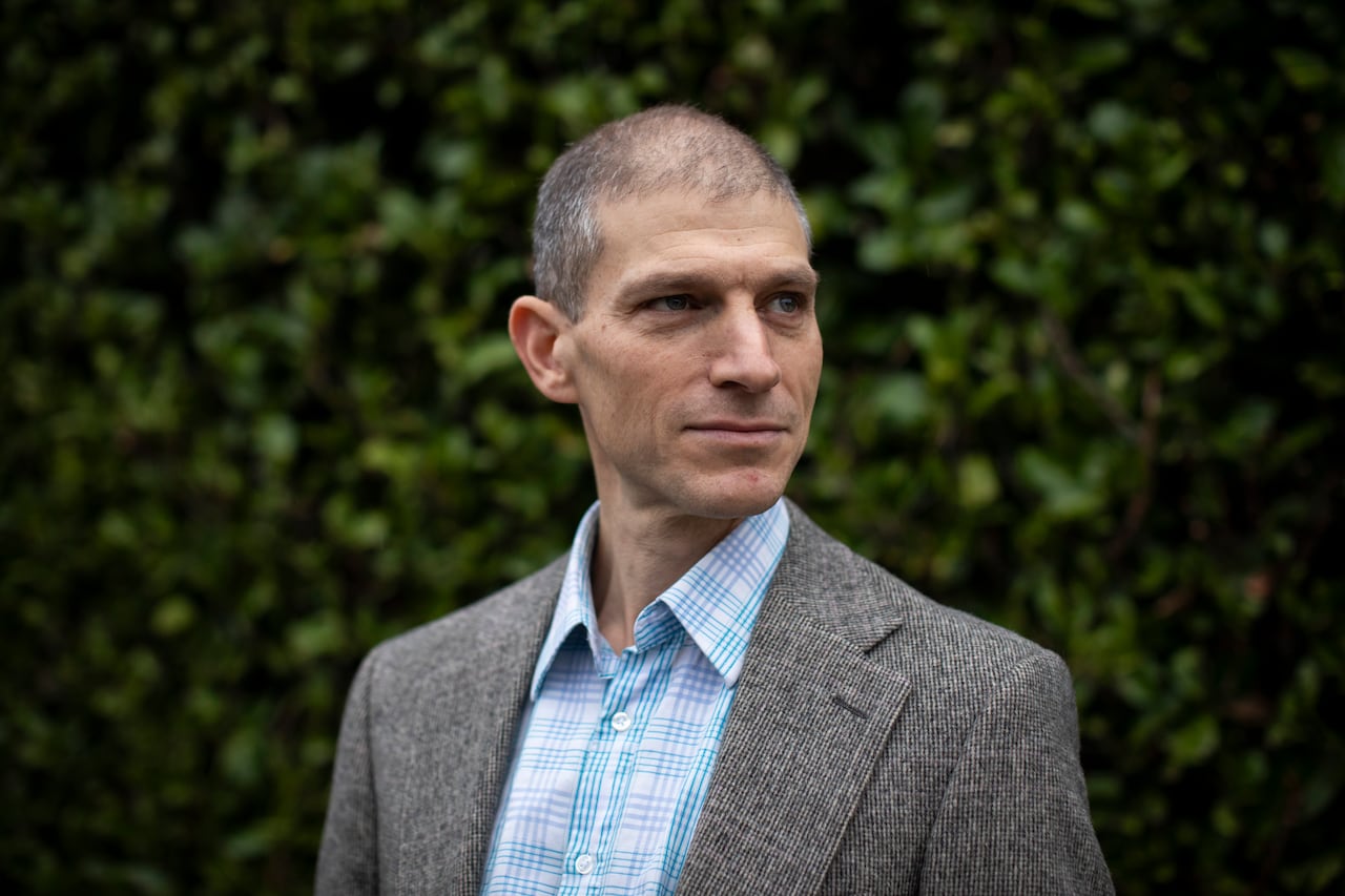 A man wearing a grey jacket and a blue checked shirt poses against a backdrop of a hedge.