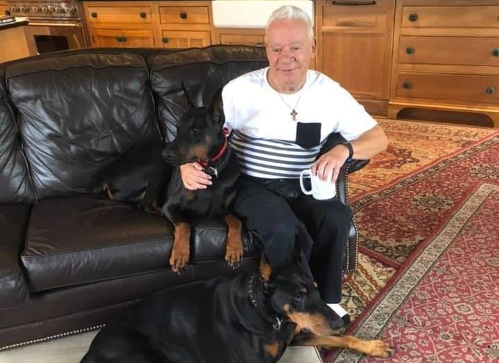 A man with white hair, wearing a white shirt with black stripes, pats one big black dog while another lies at his feet. 