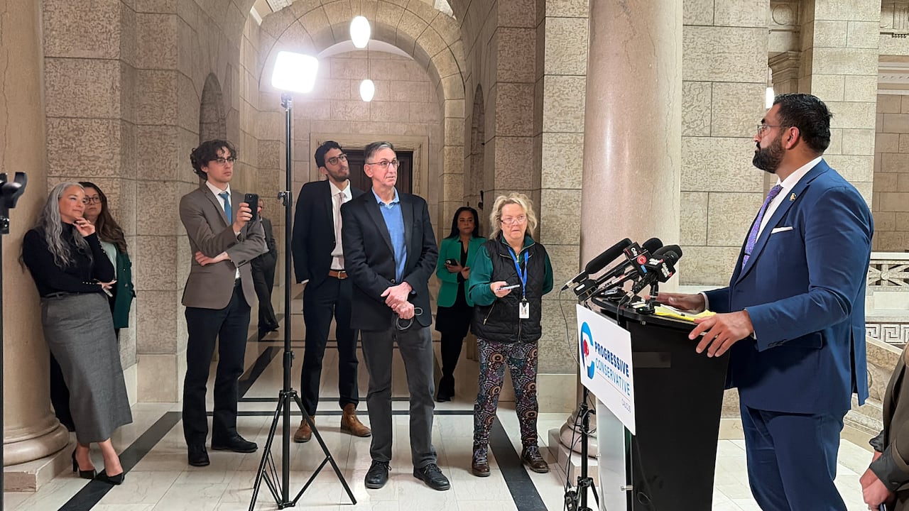 A man in a blue suit stands behind a lectern with microphones as a group of journalists ask questions.