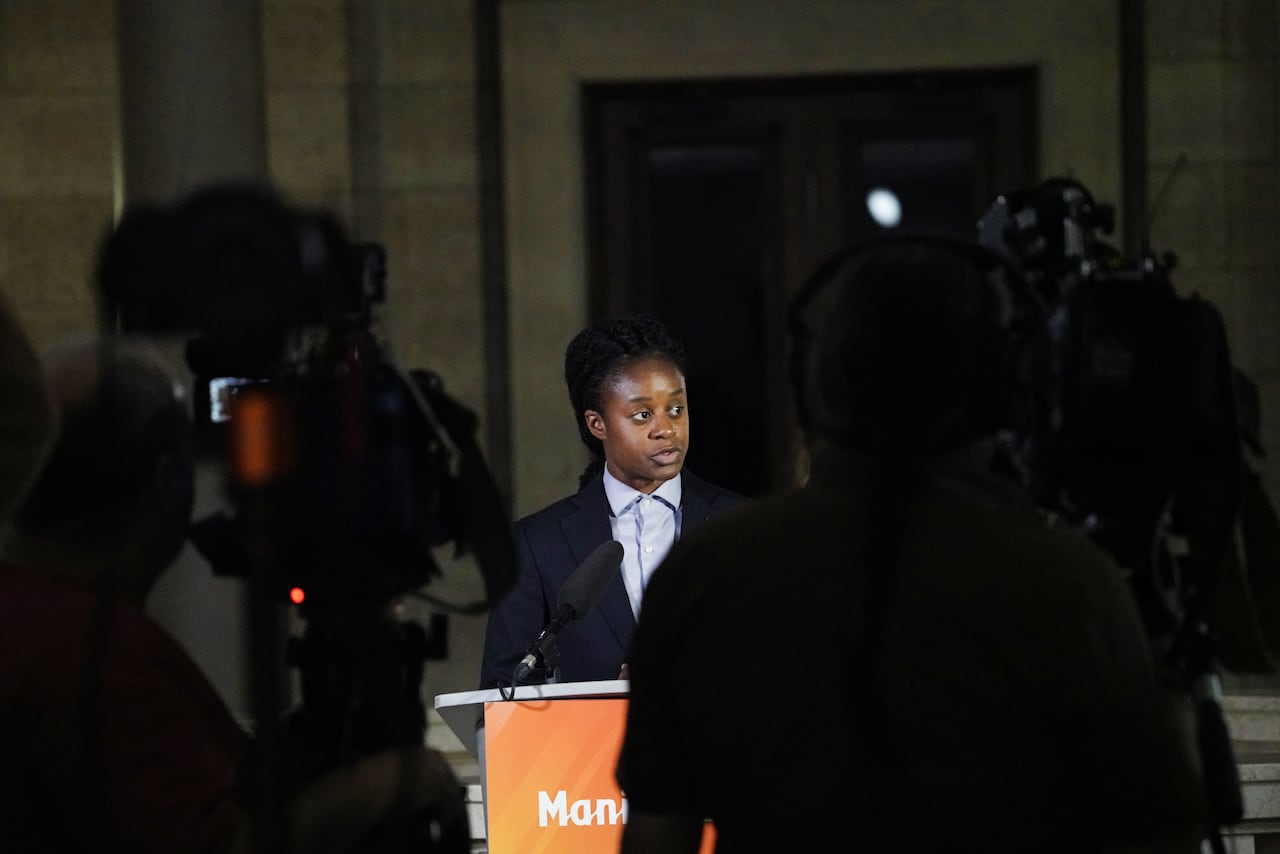 A person in a suit speaks into microphones on a lectern in front of television cameras.