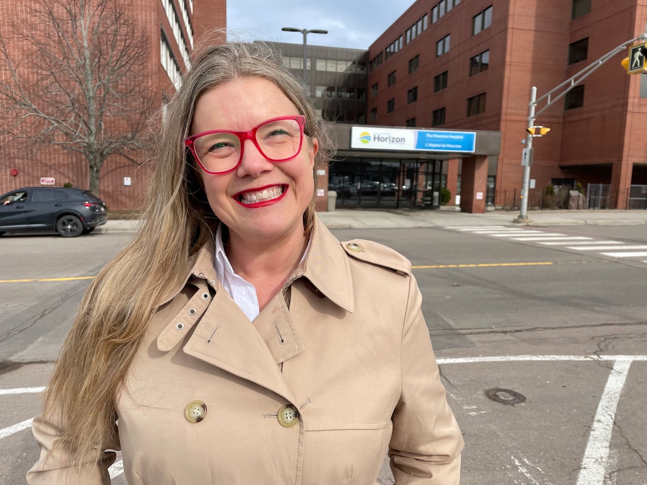 A white woman in a brown coat with long blonde hair and red glasses stands in front of the Moncton Hospital, smiling.