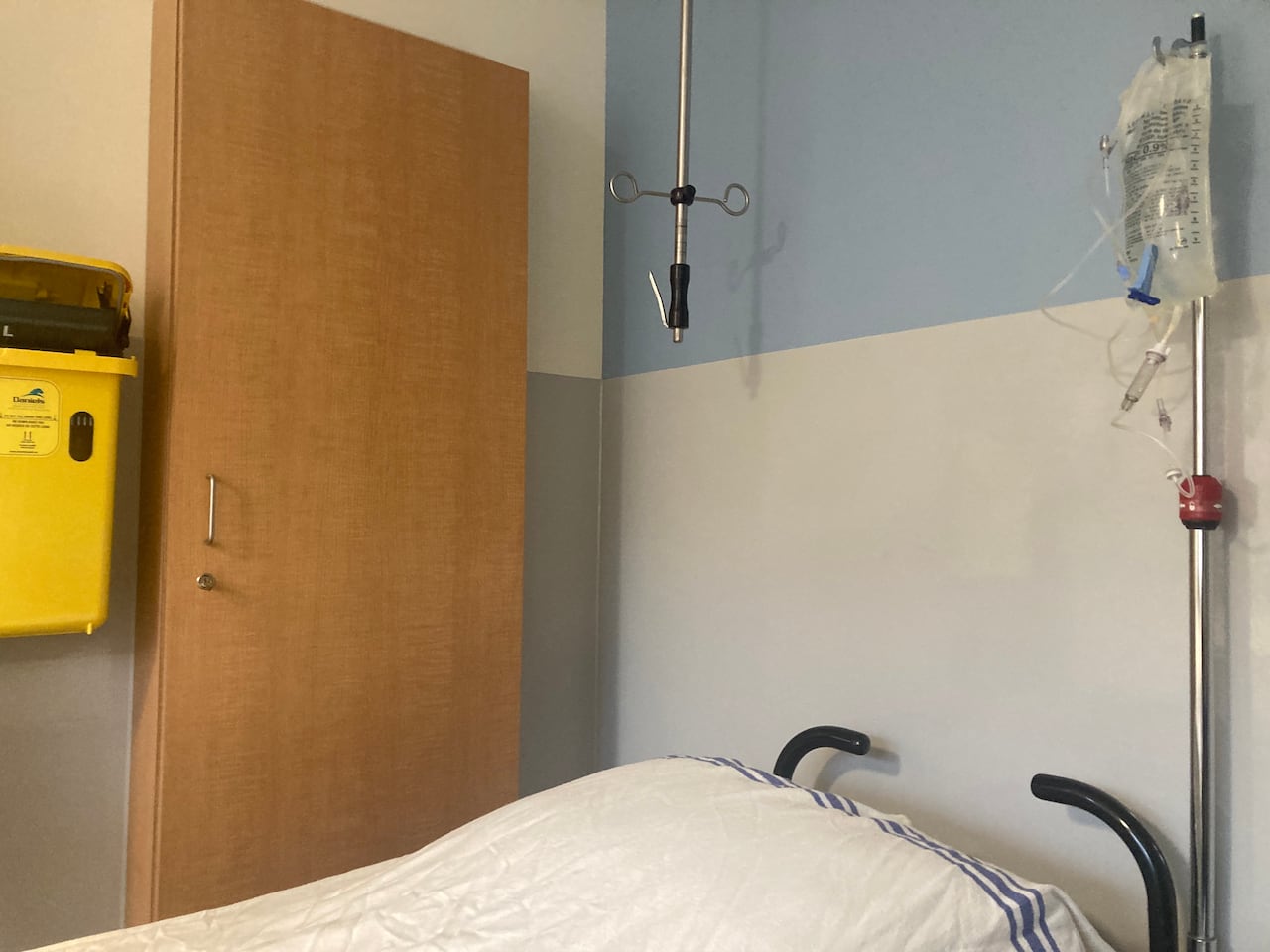 A hospital bed with white sheets, and in the background sharps disposal box and IV fluid bag.