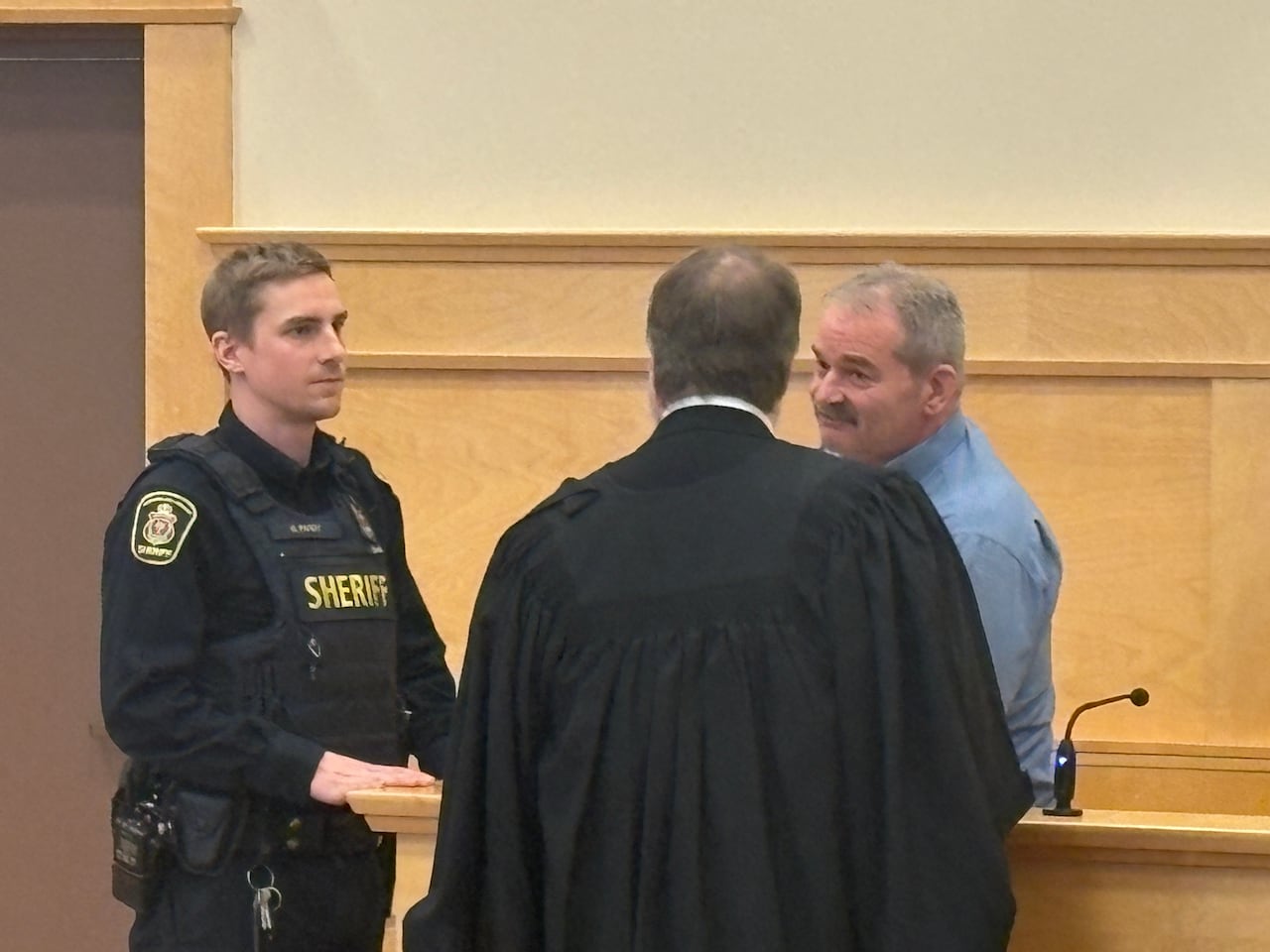 A man speaks with a lawyer while standing in a court box. A sheriff is standing with them.