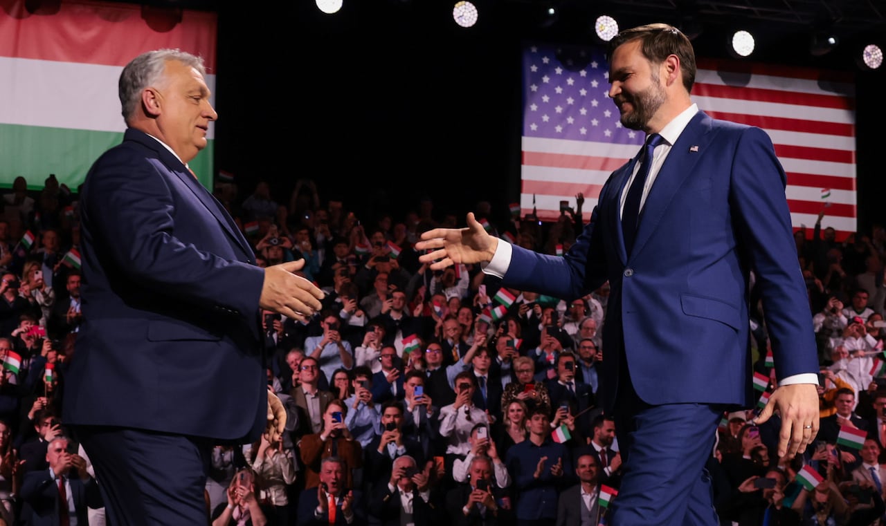 JD Vance and Viktor Orban reach out for a handshake, in front of a crowd and a backdrop of large U.S. and Hungarian flags.   