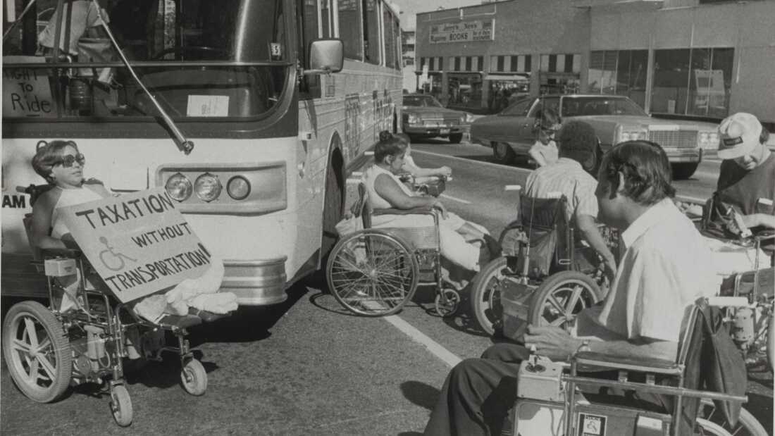 A black and white photo shows a group of seven protesters in wheelchairs surrounding a bus and stopping traffic in Denver in July 1978. A protestor holds a sign that reads: "Taxation without transportation!"