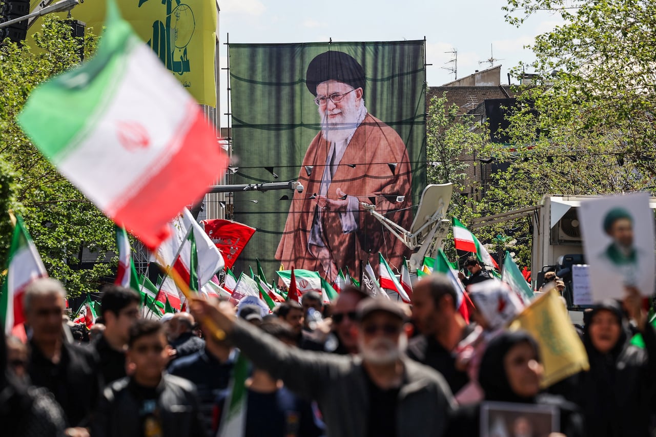 An outdoor demonstration is shown. Many people hold red and green flags, with a huge banner of a bearded man shown.