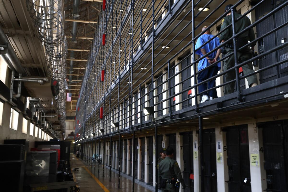 A person, seen from behind, walks with hands behind his back in front of another person next to rows of prison cells