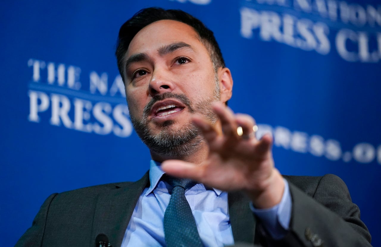 A Hispanic man speaks at a conference with his arm outstretched.