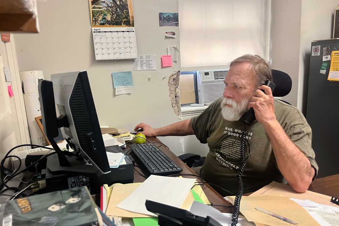 Holding a landline phone to his ear, Bill Galvin sits at an office desk with a desktop computer and keyboard on it.
