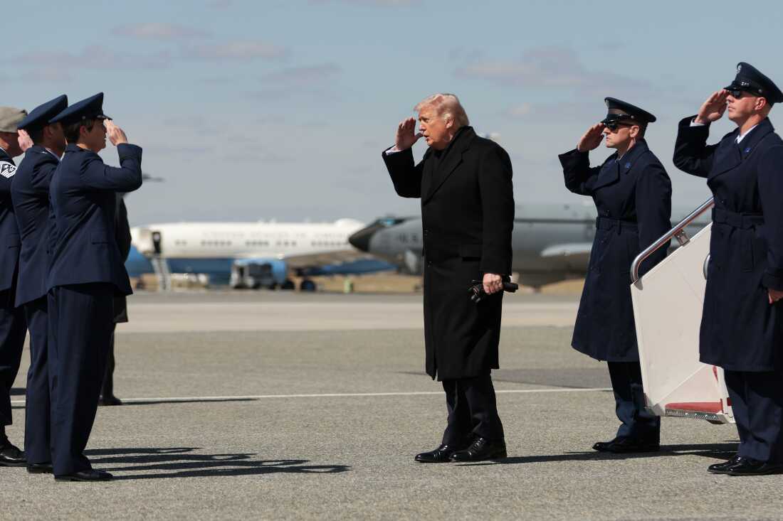 In this photo, President Trump salutes, with his hand in front his forehead, while standing and facing a short row of military personnel at Dover Air Force Base. Two airplanes are in the background.