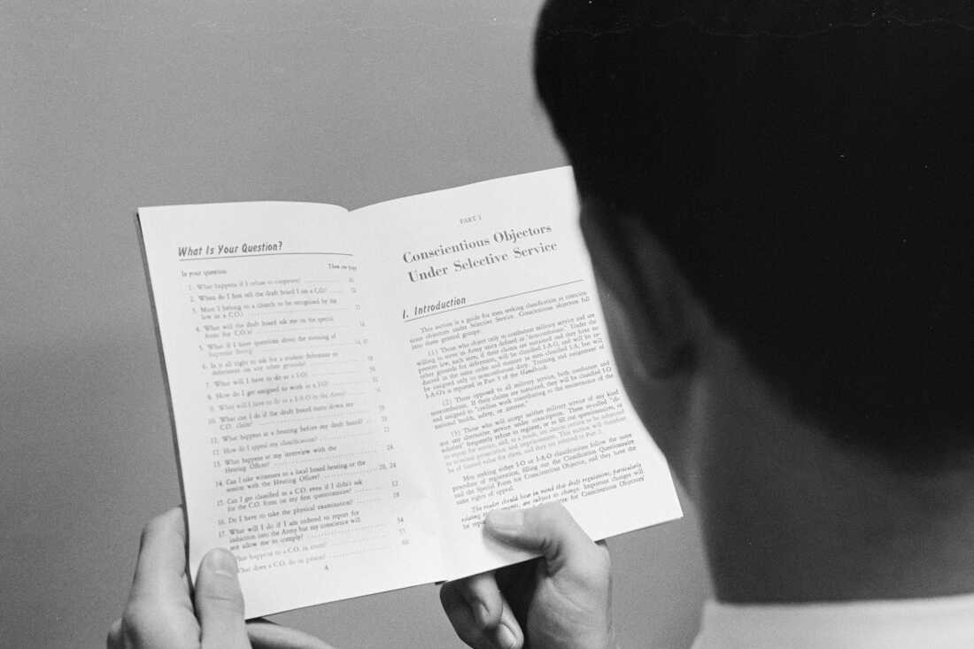 In this black-and-white photo from 1965, a man who is photographed from behind and over his left shoulder is holding a booklet that's opened to a page with the heading "Conscientious Objectors Under Selective Service."