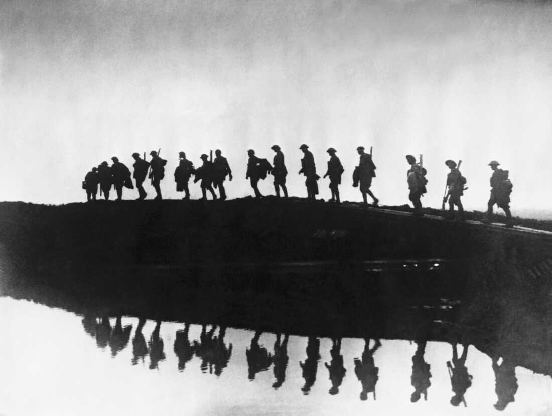A black and white photograph depicting an East Yorkshire regiment of British troops in silhouette as they negotiate their way around a rainwater-filled shell crater near the western front during World War I.