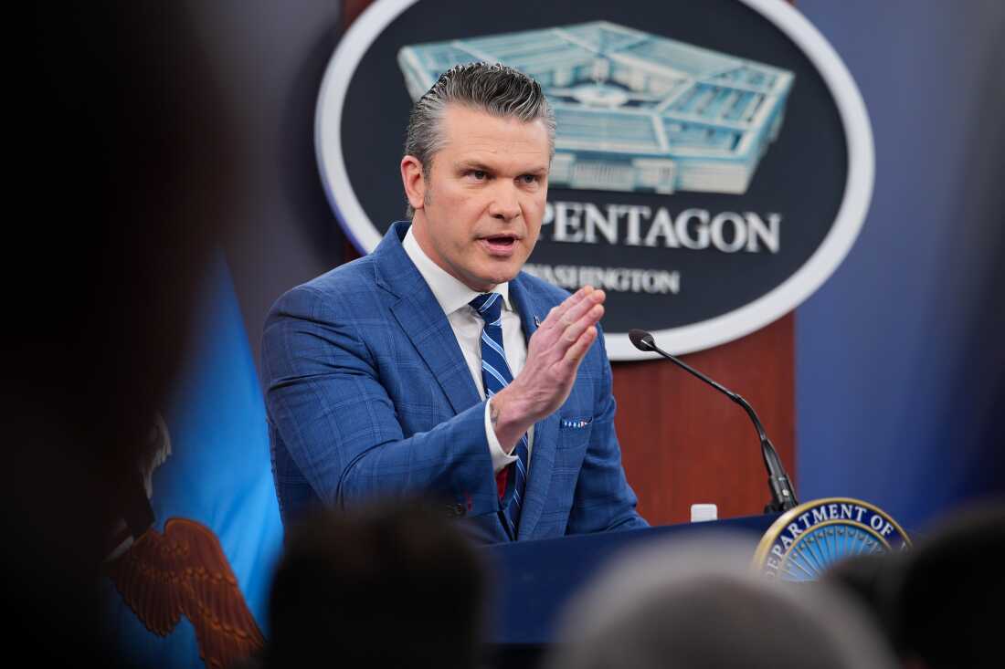 Secretary of Defense Pete Hegseth stands behind a podium  speaks during a press briefing at the Pentagon on Wednesday. He is wearing a blue suit and a blue striped tie, and is gesturing with his right hand.