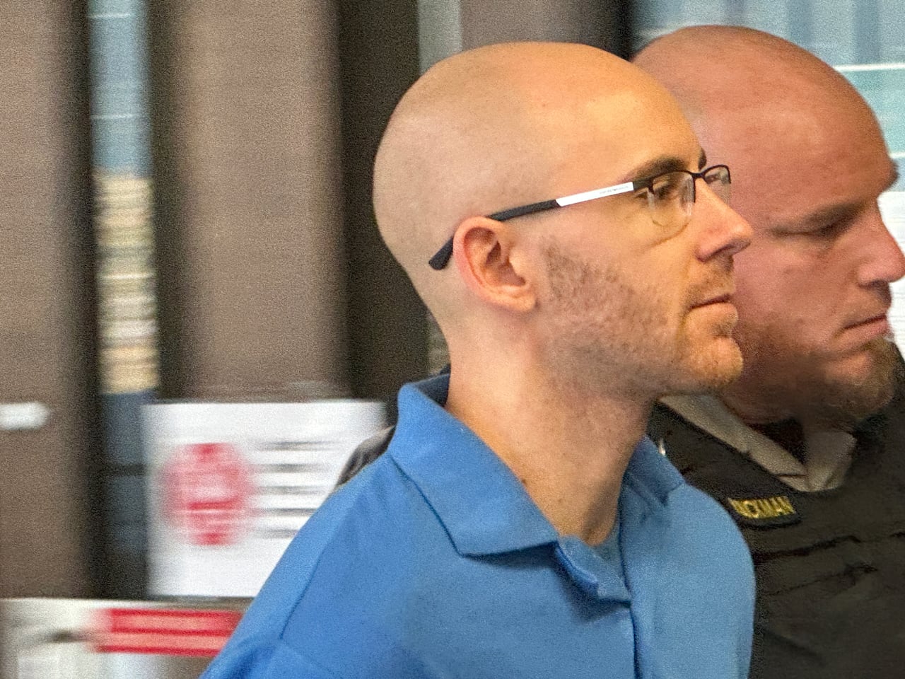 A bald white man is wearing glasses and a blue polo shirt. He's being escorted by a bald police officer.