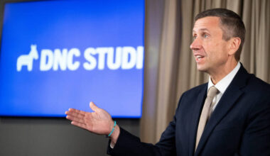 A man wearing a jacket and tie and standing in front of a screen.