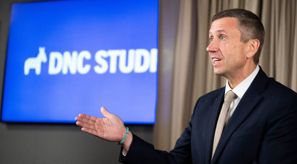 A man wearing a jacket and tie and standing in front of a screen.