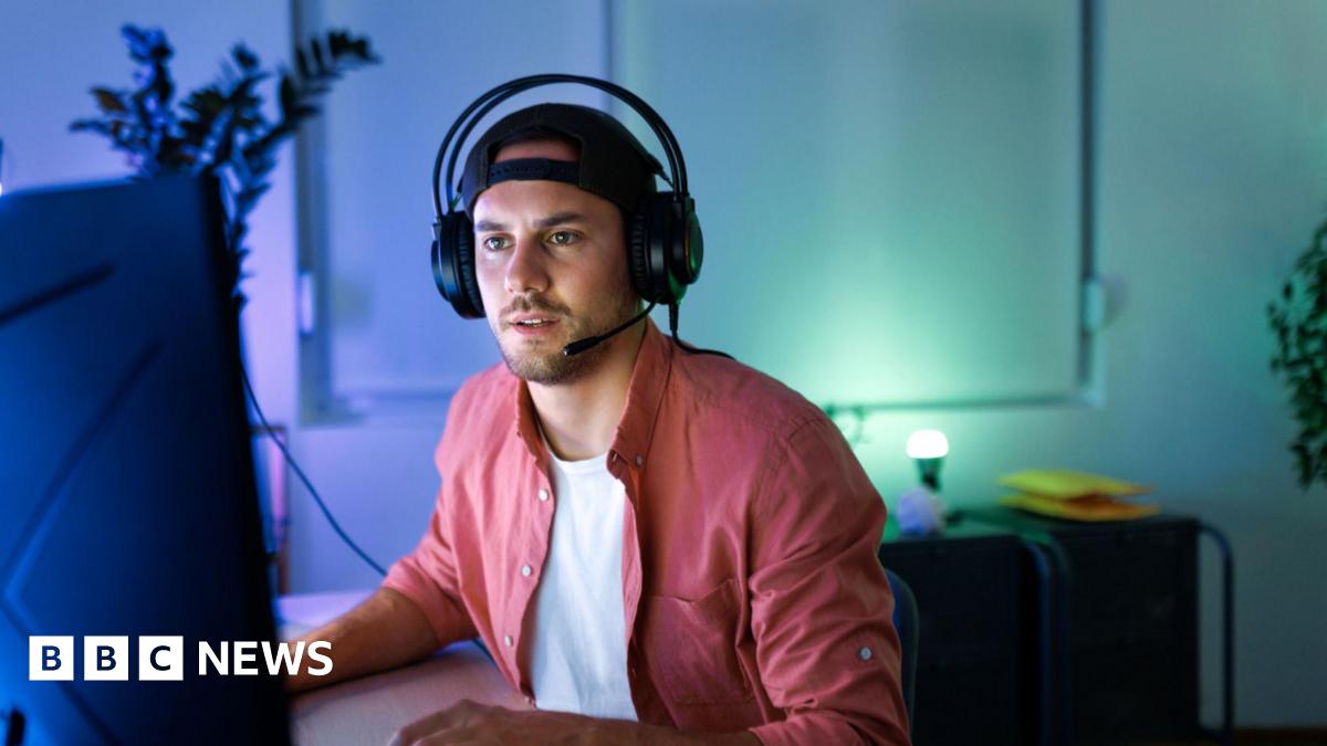 A young man in a t-shirt and open pink shirt wearing a headset with headphones and a mouthpiece is looking at a large screen with a look of concentration on his face.