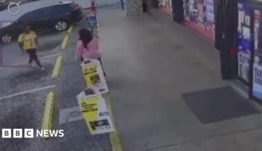 David Robinson is photographed indoors against a pale wall with a subtle floral pattern. He is shown from the shoulders up, wearing a light shirt and a dark jacket. The lighting is soft and even, and the background is uncluttered.