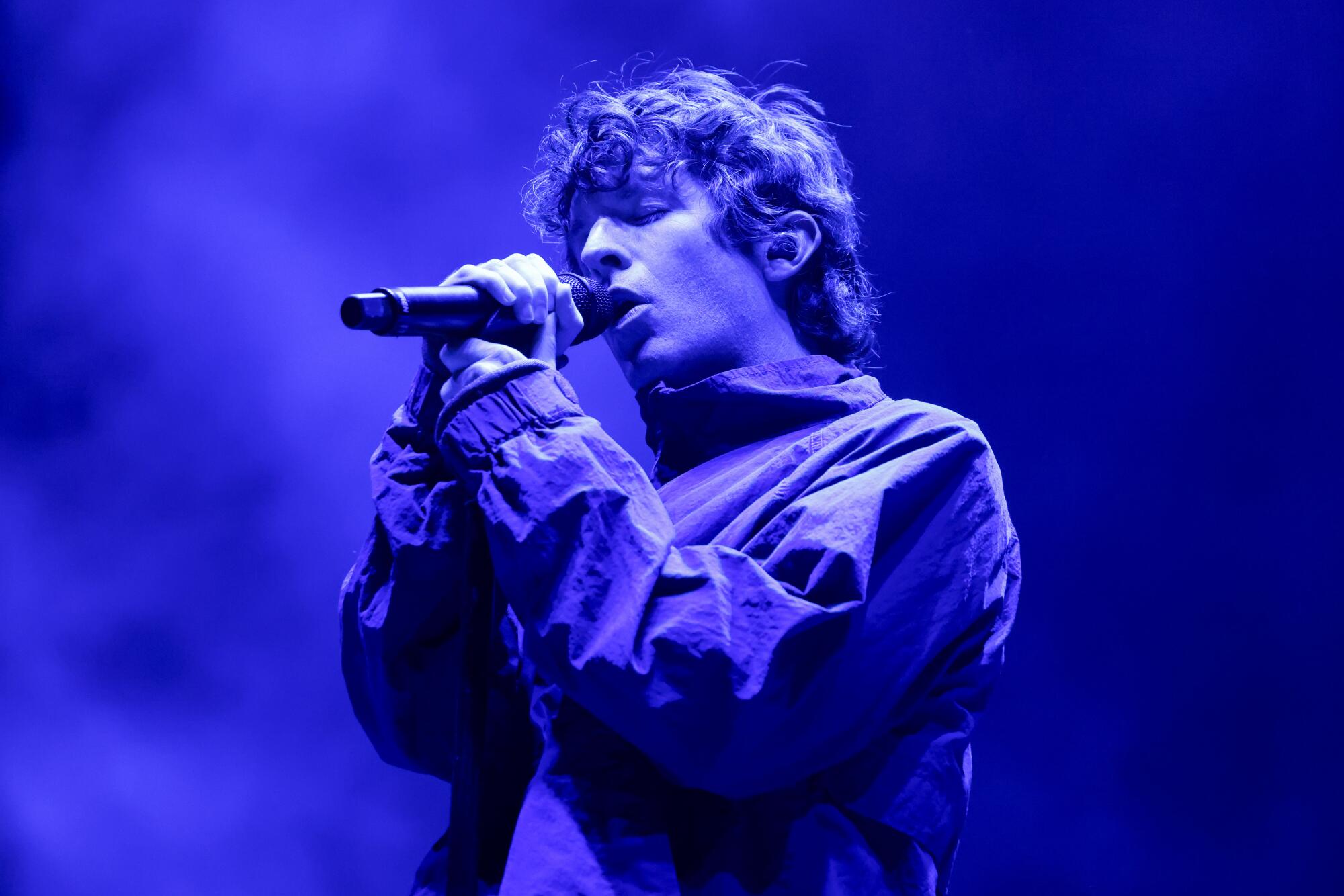 A man in a a jacket sings into a microphone while bathed in blue light