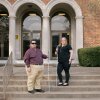 ALT TEXT: A 34 year old white man with short, black hair, dark glasses, and a plaid shirt stands to the left. A 43 year old woman with shoulder length blonde hair and an all black outfit, stands to the right. They’re both holding canes on stairs in front of a brick building.