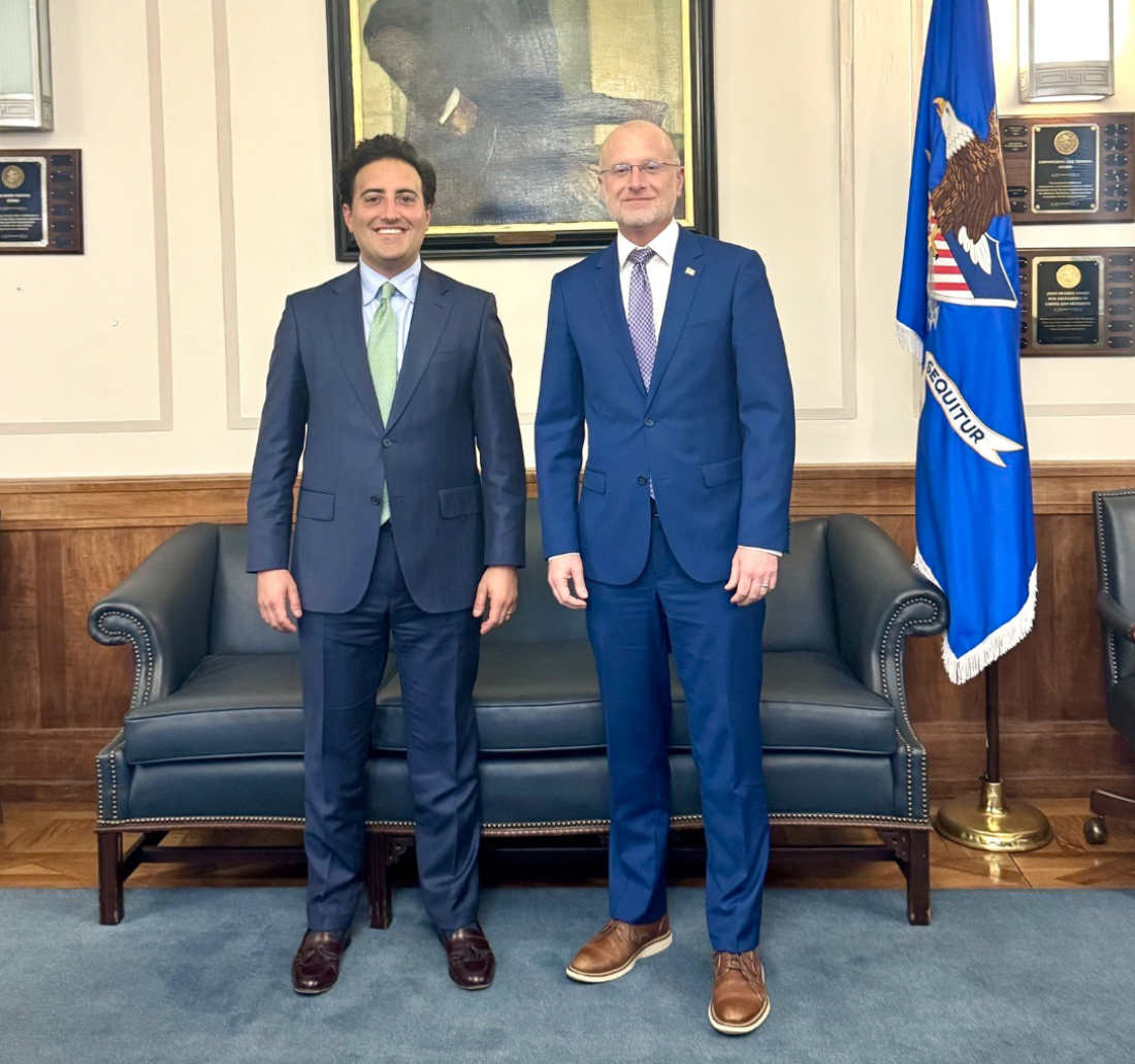 Two men dressed in suits and ties stand in front of a couch with a flag off to the side. There's a portrait on the wall behind them and plaques on the wall.