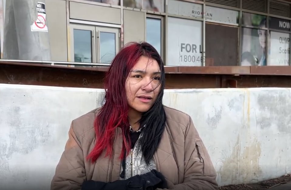 A woman stands outside wearing a few layers. Her red and black hair blows in the wind.