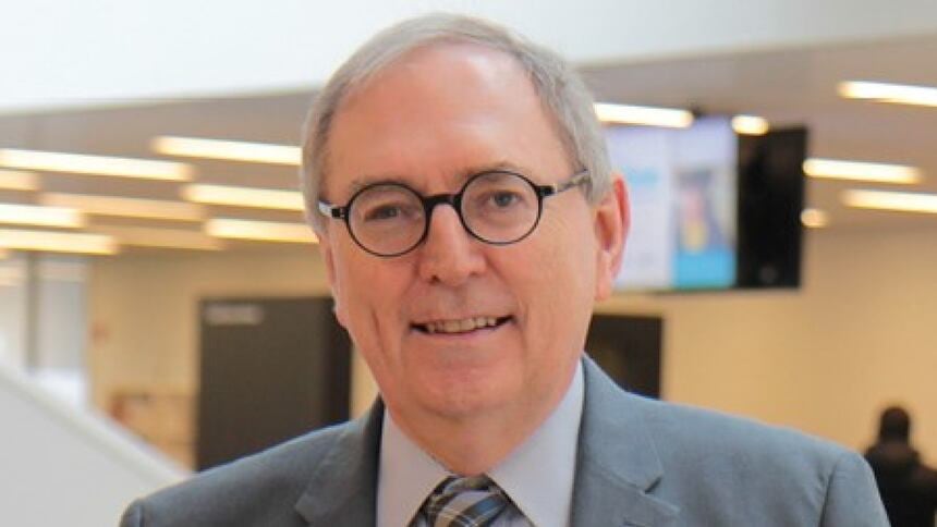 A man with a grey suit and tie and black rimmed glasses smiles at the camera. 