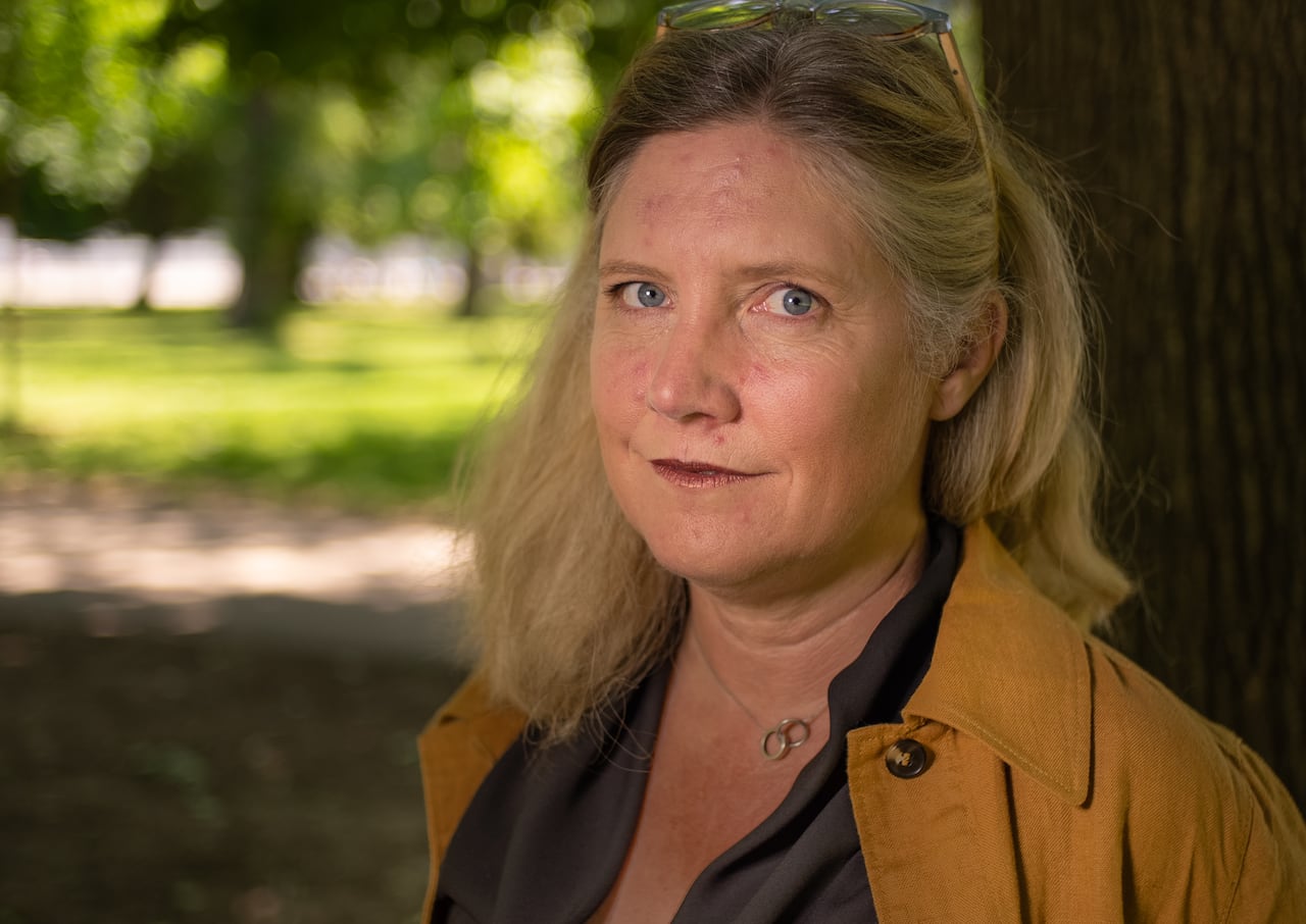 A woman with shoulder length blond hair and wearing a sand coloured jacket over a dark top stands outdoors in a park area.