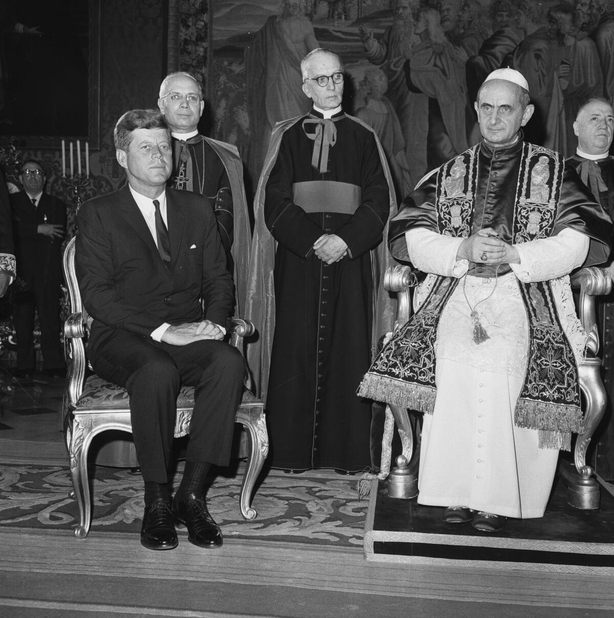 A black-and-white photo of a man in dark suit and tie seated next to a man in ornate religious vestments and a white skullcap