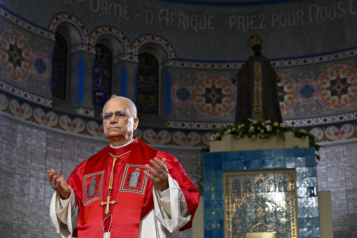 A man with glasses, in red vestments, holds out his hands in prayer in a room with ornate blue and yellow mosaic walls