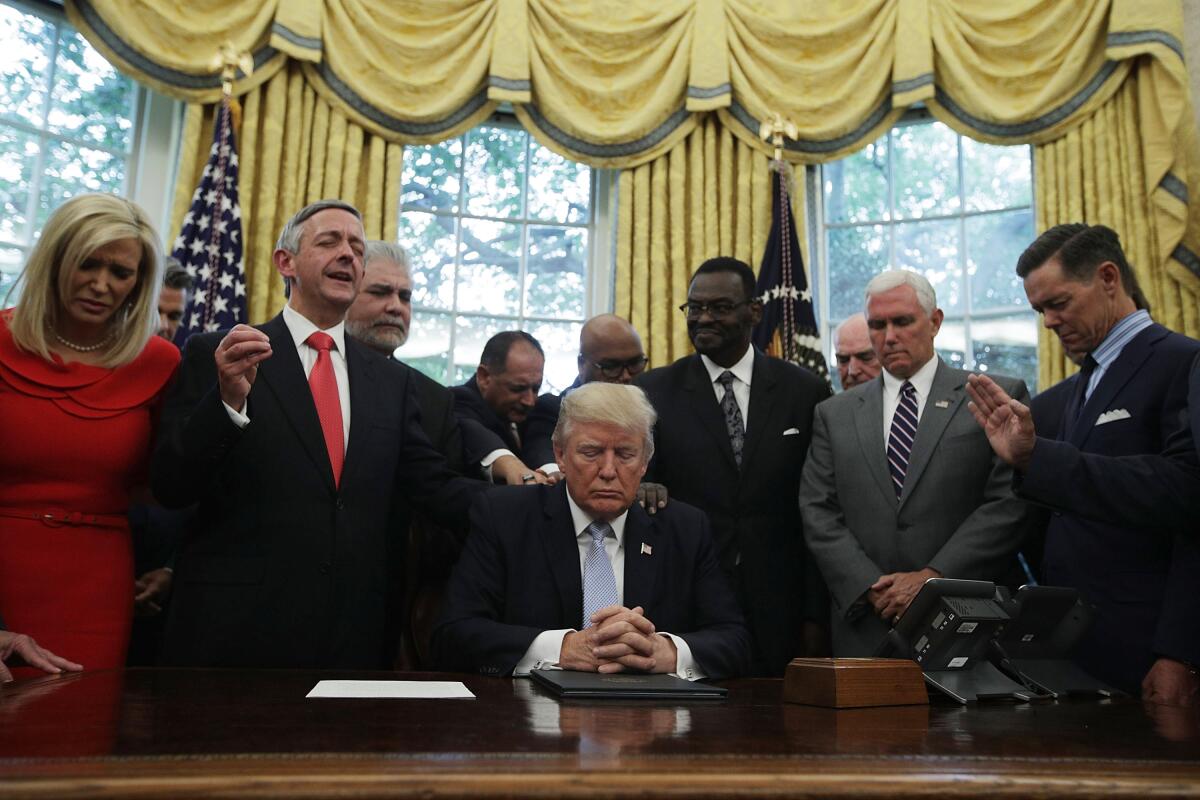 A man in a dark suit, hands clasped on a desk, is surrounded by other people standing near windows with gold curtains