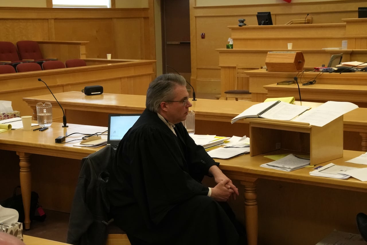 A man wearing a black robe sits at a desk in a courtroom.