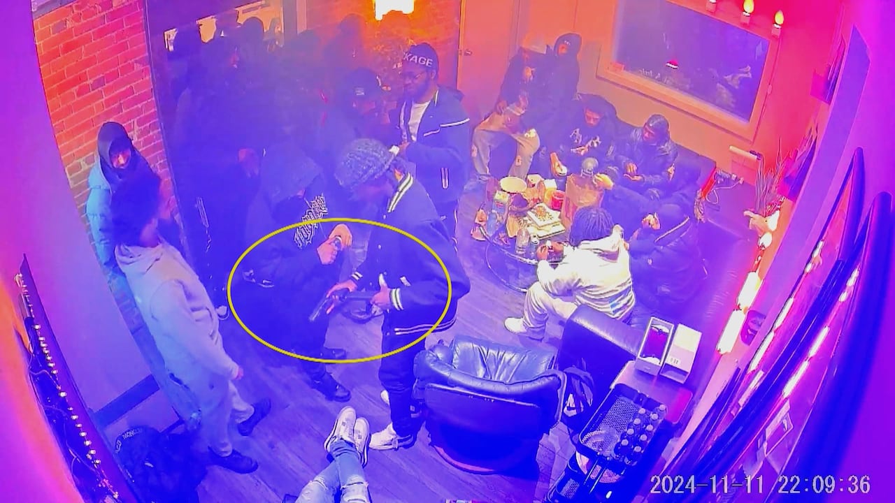 A group of people are seen on surveillance video. A circle highlights one man loading a gun