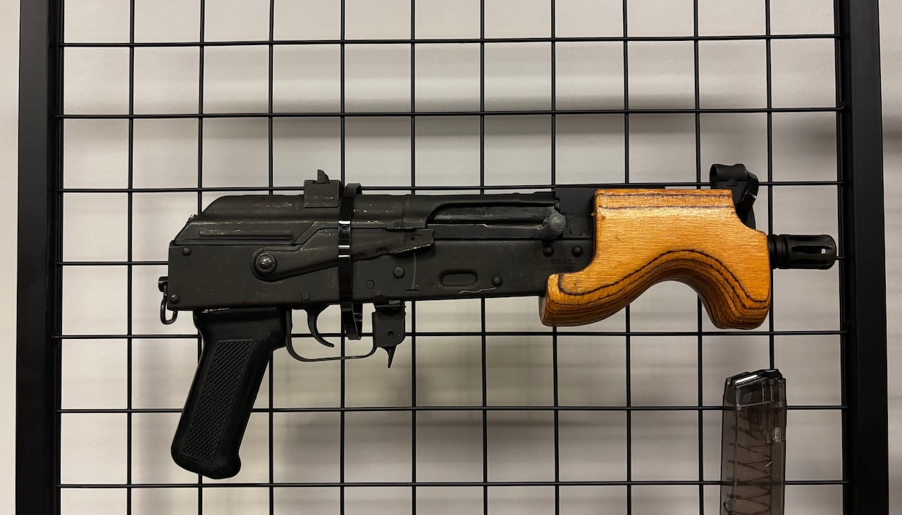A black gun with a wooden hand guard is zip tied to a black metal grid.