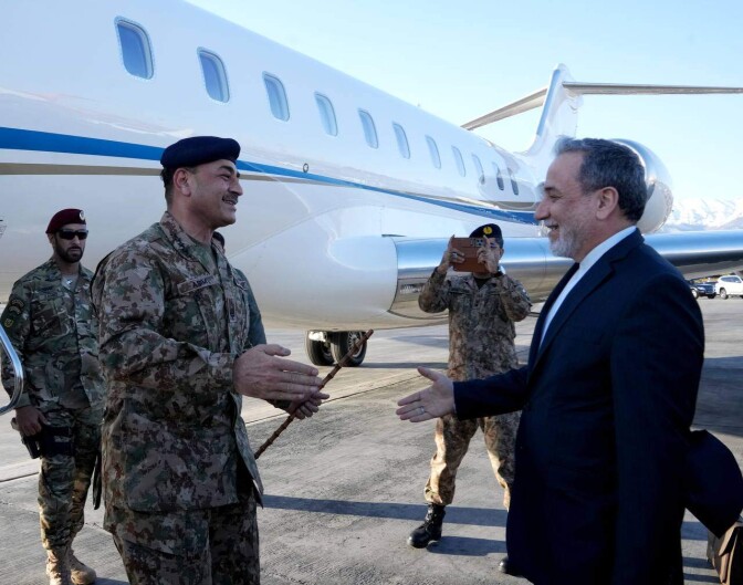 A man in army fatigues greets a man in a dark suit on the tarmac in front of a jet.