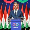 Peter Magyar stands behind a lectern with two microphones attached to it as he speaks at a Monday press conference. Multiple Hungarian flags are standing in a row behind him, and the front of the lectern says "Tisza" on it.