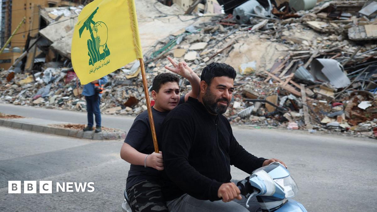 Two people ride on a moped, while one holds a Hezbollah flag and holds two fingers in the air, on a street in Beirut in front of a huge pile of rubble where a building once stood, taken on Friday.