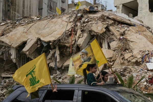 Displaced residents wave Hezbollah flags, including one bearing a picture of its leader, Naim Qassem, as they pass rubble of destroyed buildings in Dahiyeh, Beirut's southern suburbs, Lebanon, Friday, April 17, 2026, following a ceasefire between Israel and Hezbollah. (AP Photo/Bilal Hussein)