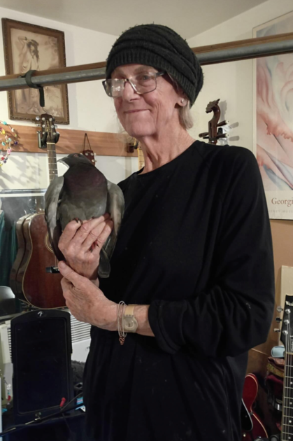An older person in a black outfit and hat gently holds a large pigeon, surrounded by guitars and framed art in a cozy room.