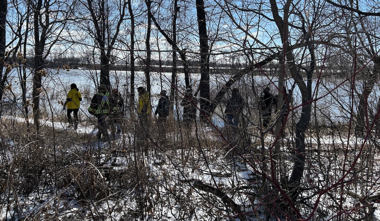 A line of people wearing winter jackets is seen walking on a path through a wooded area on a snowy day