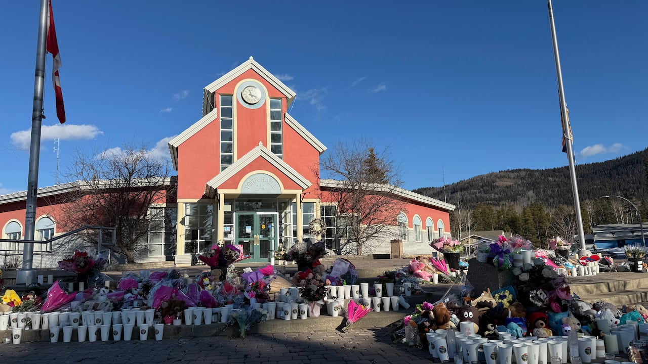 Dozens of bunches of flowers and candles in cups on the ground in front of an orange town hall building.