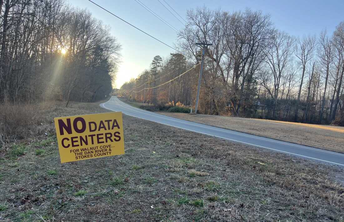 A yellow sign saying "NO DATA CENTERS for Walnut Cove... the Dan River & Stokes County" is staked into the ground by the side of a rural road with dried grass and leafless trees on each side.