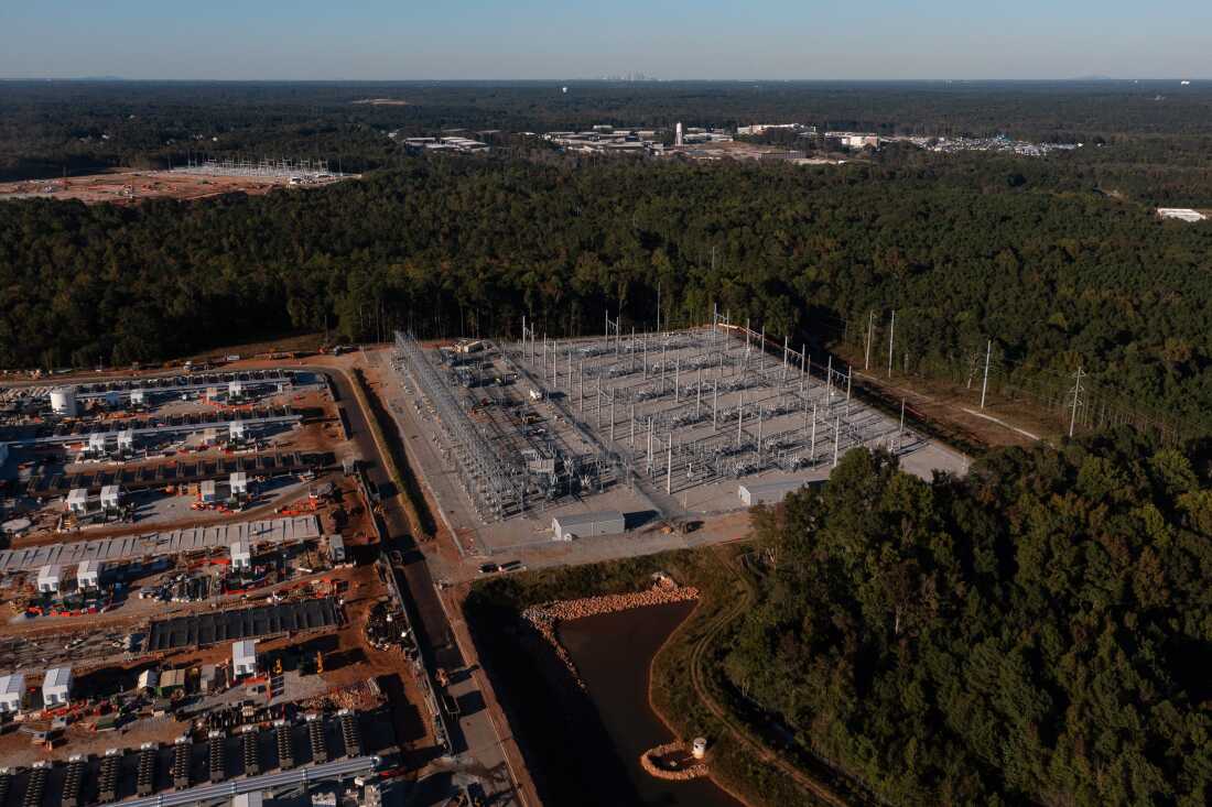 In this 2024 aerial photo, a QTS data center complex on the left side of the frame is under construction in Fayetteville, Georgia. A power substation stands adjacent to it, on the right side of the frame. Green trees surround both sites.