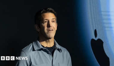 John Ternus standing on stage in front of an Apple logo. He is wearing a grey shirt.